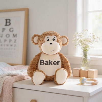 A brown stuffed monkey toy with the name "Baker" on its chest sits on a white dresser next to a vase of white flowers and a wooden block.