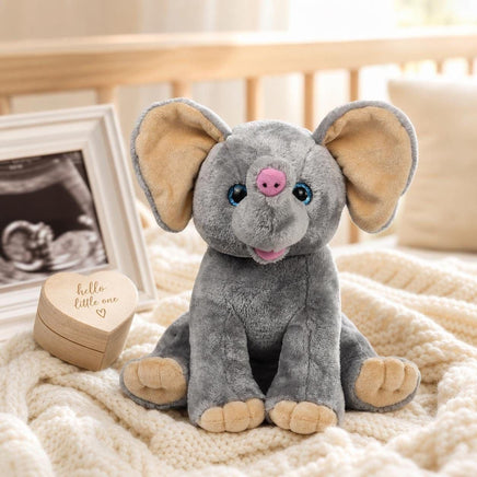 A gray stuffed elephant toy sits on a white blanket, with a wooden box and a framed photograph of a baby in the background.
