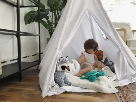 A young child sits inside a white teepee tent, surrounded by stuffed animals, in a cozy indoor setting with a potted plant in the background.