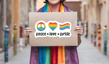 A person is holding a sign with the words "Peace, love, pride" written on it, along with a peace symbol and a rainbow flag.