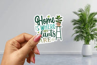 A hand holding a sticker with the text "Home is where my plants are" against a backdrop of a potted plant and a gray wall.