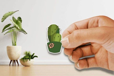 A hand is holding a sticker of a cactus plant on a wall, with a potted plant and a wooden floor in the background.