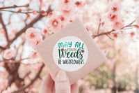 A hand holding a small square object with a sticker on it, against a background of pink flowers.