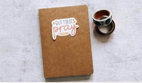 A brown notebook with a sticker that says "BUT FIRST PRAY" is placed on a white surface next to a roll of tape.