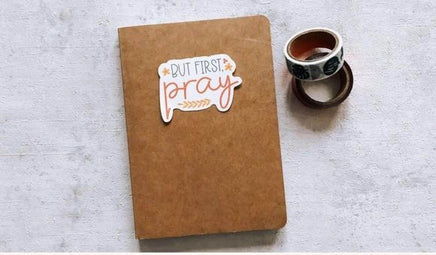 A brown notebook with a sticker that says "BUT FIRST PRAY" is placed on a white surface next to a roll of tape.