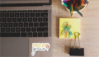 a laptop with a sticker that says "SAVED BY GRACE", a yellow sticky note with colorful pencils, and a black binder clip.