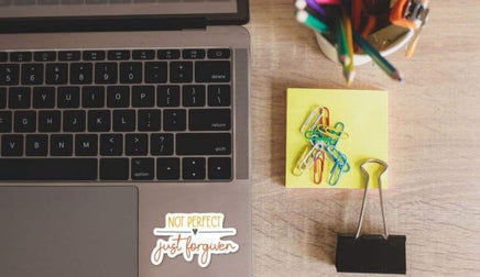 a laptop, a yellow sticky note, a binder clip, and a cup of colored pencils on a wooden surface.