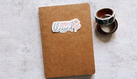 A brown notebook with a sticker that says "Simply Blessed" is placed on a white surface next to a roll of tape.