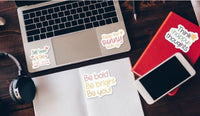 a laptop, a notebook, a smartphone, and some stickers on a wooden surface.