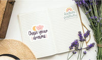 an open notebook with a sticker that says "Chase your dreams" and another sticker that says "Radiate positivity". The notebook is placed on a white surface, with a hat and some purple flowers nearby.