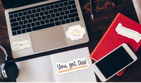 a laptop, a smartphone, and some books arranged on a wooden surface. The laptop has stickers on it, and there is a red book and a notebook with a sticker on it as well. The smartphone is placed next to the notebook, and there is a pair of headphones nearby. The overall scene conveys a sense of productivity and organization.