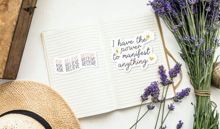 an open notebook with a motivational quote, a bouquet of purple flowers, and a straw hat on a white surface.
