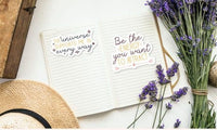 a notebook with a motivational quote, a bouquet of purple flowers, and a straw hat, all arranged on a white surface.