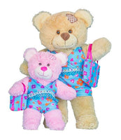 Two teddy bears, one light brown and one pink, are standing next to each other. The light brown teddy bear is wearing a blue dress with a floral pattern, and the pink teddy bear is holding a blue and pink gift bag.