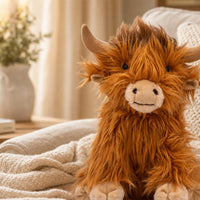 A plush toy of a furry, orange-brown animal resembling a yak or a similar creature is sitting on a bed, with a white vase and a plant in the background.