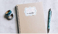 a spiral notebook with a sticker that says "COFFEE IS MY FAVORITE CO-WORKERER", a pen, and some tape rolls on a white surface.
