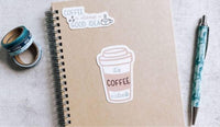 a spiral notebook with a coffee cup sticker on it, along with a pen, a pen roller, and some tape rolls.