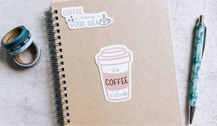 a spiral notebook with a coffee cup sticker on it, along with a pen, a pen roller, and some tape rolls.
