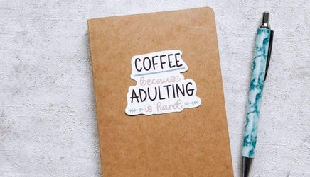 A brown notebook with a sticker that says "COFFEE BECAUSE ADULTING IS HARD" next to a pen on a white surface.