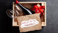 A wooden box containing a bottle of wine, two wine glasses, and a gift box with a red ribbon. There is also a brown paper with a red ribbon and a heart-shaped tag that reads "Will you be my Valentine?".