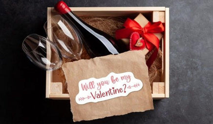 A wooden box containing a bottle of wine, two wine glasses, and a gift box with a red ribbon. There is also a brown paper with a red ribbon and a heart-shaped tag that reads "Will you be my Valentine?".
