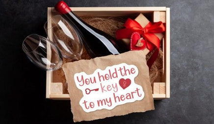 A wooden box containing a bottle of wine, two wine glasses, and a gift box with a red ribbon. There is also a handwritten note that reads "You hold the key to my heart".