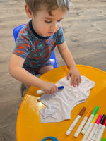 A young child is sitting at a table, intently coloring a white shirt with a blue marker.