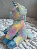 A plush toy of a unicorn with a blue horn and multicolored fur is sitting on a white bedspread.