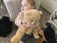 A young child is sitting on the floor, holding a large teddy bear in their arms. There are also two smaller stuffed animals, one black and one brown, in the background.