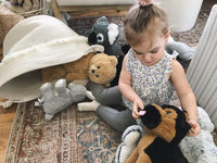 A young girl sits on the floor surrounded by various stuffed animals, including a teddy bear, a dog, and a cat.