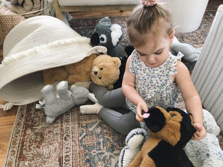 A young girl sits on the floor surrounded by various stuffed animals, including a teddy bear, a dog, and a cat.