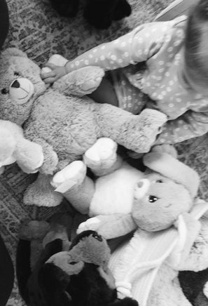 A child is sitting on the floor surrounded by various stuffed animals, including a large teddy bear and a smaller bunny.
