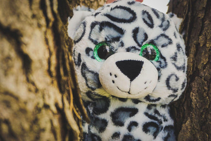 A stuffed toy leopard with large, green eyes peeking out from behind a tree trunk.