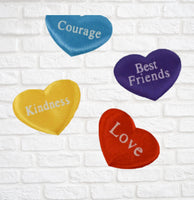 four heart-shaped objects with different messages written on them, placed against a white brick wall.