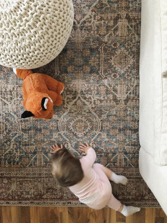 A young child in a pink outfit is reaching for a stuffed animal on a patterned rug, with a white ottoman and a white couch visible in the background.