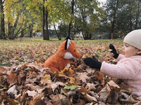 A young child in a pink jacket and hat is playing with a large orange stuffed animal fox in a park filled with fallen autumn leaves.
