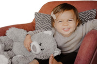 A young girl sits on a red chair, smiling and holding a large gray teddy bear.