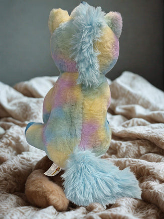 A colorful plush toy, resembling a unicorn or a teddy bear, is sitting on a bed with a white and beige patterned blanket.