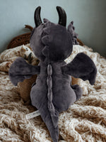 A plush toy dragon with black horns and wings sits on a bed, with a small stuffed animal cat visible in the background.