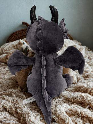 A plush toy dragon with black horns and wings sits on a bed, with a small stuffed animal cat visible in the background.