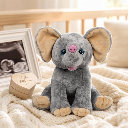 A gray stuffed elephant toy sits on a white blanket, with a wooden box and a framed photo of a baby in the background.