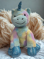 A plush toy of a unicorn with a blue horn and glittery feet is sitting on a white surface.