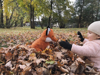 A young child in a pink jacket and hat is playing with a stuffed orange fox in a park filled with fallen autumn leaves.