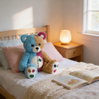 A blue and brown teddy bear sits on a bed, with an open book and a lamp nearby.