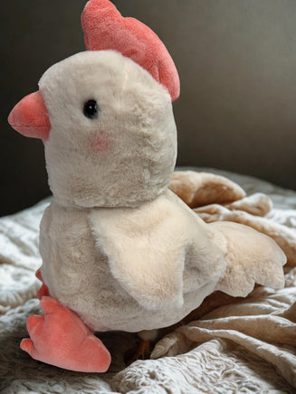 A plush toy of a chicken with pink accents is sitting on a bed.