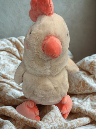 A plush toy of a chicken with a pink beak and feet is sitting on a patterned fabric.