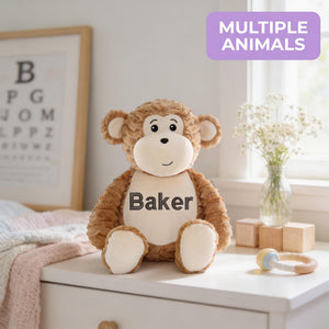 A brown stuffed monkey toy with the name "Baker" printed on it, sitting on a white dresser next to a vase of flowers and a baby rattle.