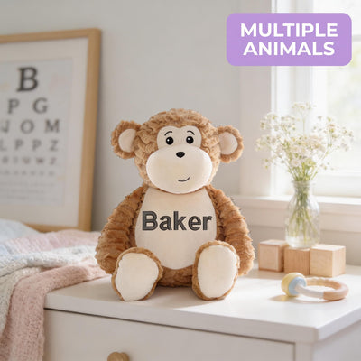 A brown stuffed monkey toy with the name "Baker" printed on it, sitting on a white dresser next to a vase of flowers and a baby rattle.