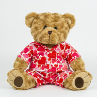 A teddy bear wearing a red and white heart-patterned pajama top and matching pajama bottoms sits on a white background.