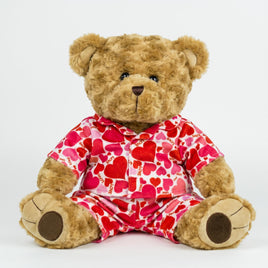 A teddy bear wearing a red and white heart-patterned pajama top and matching pajama bottoms sits on a white background.